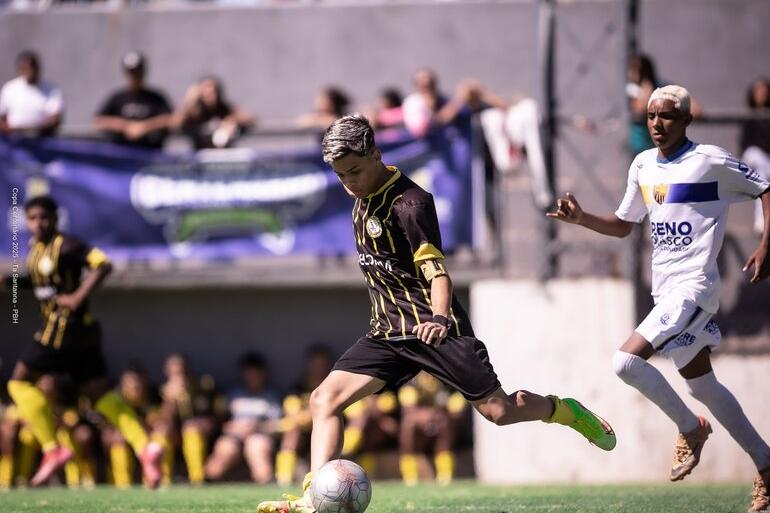 Copa Centenário movimenta campos de BH neste final de semana com jogos em três categorias Em um campo de futebol society, um jovem atleta com uniforme preto e amarelo prepara o chute com a perna esquerda, enquanto a bola está à sua frente. Ao lado, um jogador de uniforme branco e azul acompanha a jogada. Ao fundo, desfocados, aparecem outros atletas, integrantes das equipes e parte da arquibancada. A partida acontece durante o dia, sob forte iluminação solar.
