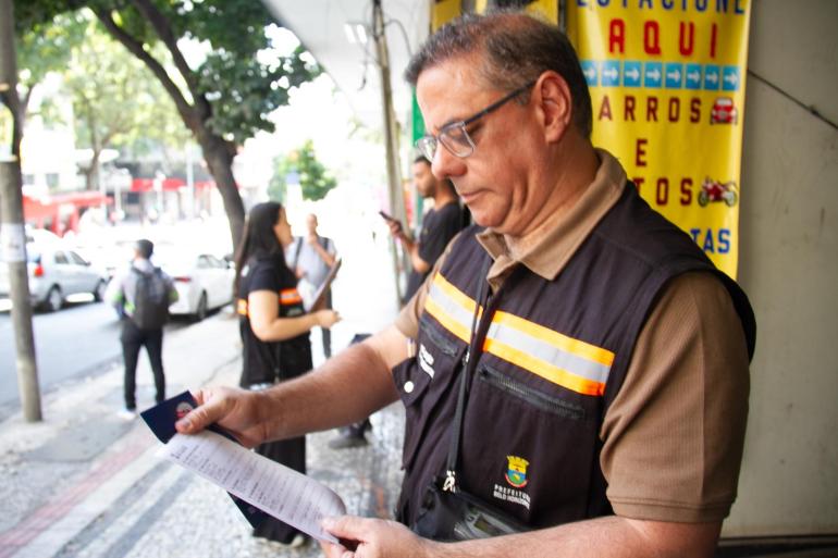 PBH intensifica fiscalização e quase 3 toneladas de lixo são retiradas do Centro e Lagoinha em apenas um dia Fiscal de colete da Prefeitura de Belo Horizonte analisa um documento impresso na calçada durante ação de fiscalização de poluição visual na cidade.