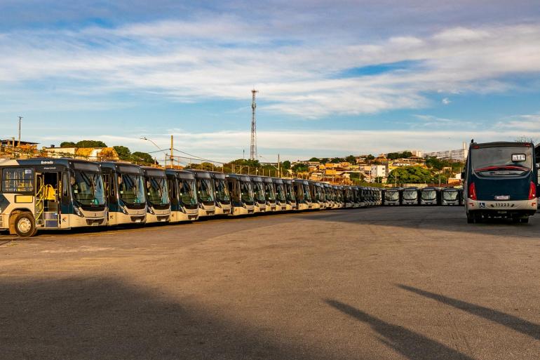 Transporte coletivo de BH recebeu 219 novos ônibus neste ano, renovando a frota para mais conforto e segurança dos usuários A foto mostra uma sequência de ônibus urbanos estacionados lado a lado em um amplo pátio asfaltado. Os veículos, predominantemente nas cores bege e preto, estão alinhados em perspectiva, formando uma longa fileira. À direita, um ônibus aparece de costas, em primeiro plano. Ao fundo, há construções, áreas verdes, postes e uma torre de telecomunicações. O céu está parcialmente nublado, com tons de azul e branco.