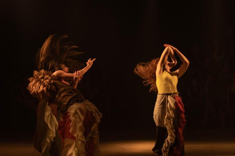 Em um palco escuro com iluminação amarelada, três bailarinas realizam um movimento de dança expressivo. À esquerda, duas estão curvadas, sendo que uma delas tem os cabelos jogados para cima em função do movimento. À direita, a terceira bailarina está em pé, com os braços erguidos sobre a cabeça e o cabelo cacheado em movimento para trás. As três vestem regatas e calças amplas com babados nas cores creme e vinho.