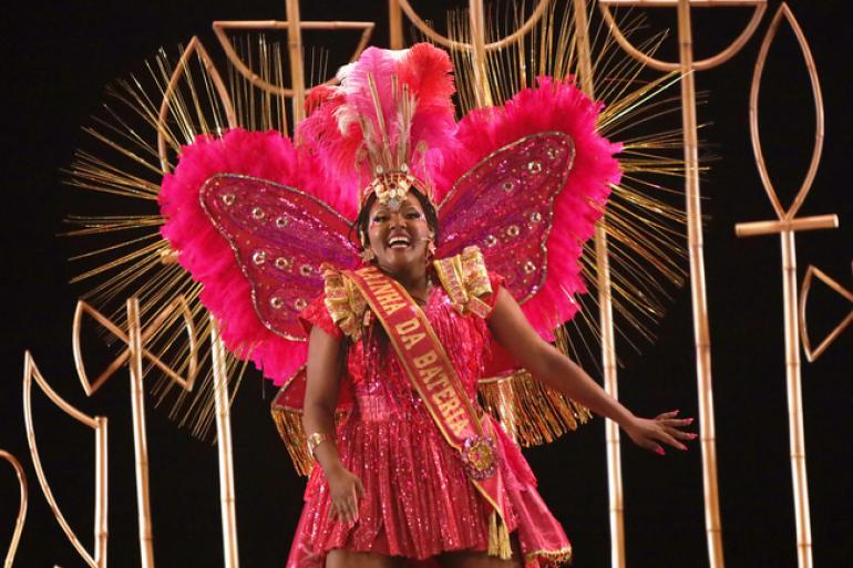  Fotografia em plano médio, no palco do espetáculo “Aída: A Rainha da Bateria”, da Cyntilante Produções. No centro, a atriz, uma mulher negra, sorri radiante enquanto dança com os braços abertos. Ela veste a fantasia de Rainha da Bateria: um vestido curto rosa-choque brilhante com detalhes dourados e uma faixa transversal com o escrito "RAINHA DA BATERIA".