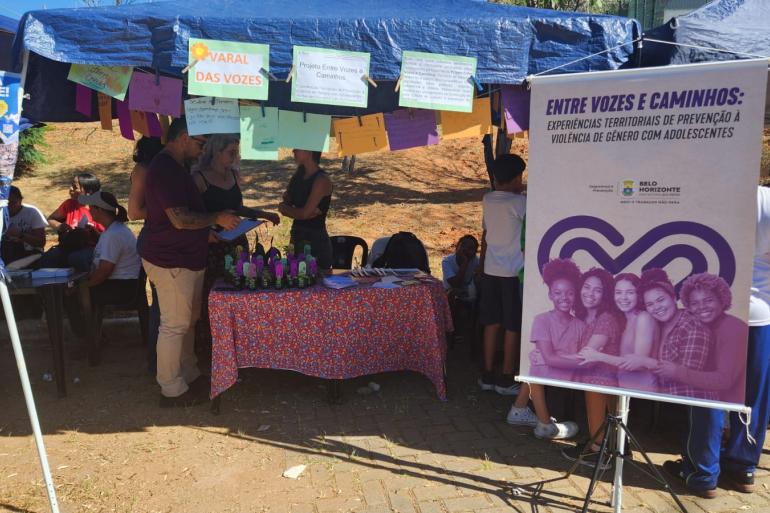 Projeto Entre Vozes e Caminhos fortalece a prevenção à violência de gênero entre adolescentes Foto de um estande montado ao ar livre, sob tendas azuis, com uma mesa coberta por uma toalha colorida, com materiais informativos. Cartazes coloridos estão pendurados em um varal, com mensagens relacionadas ao projeto Entre Vozes e Caminhos. À direita, um grande banner traz o título “Entre Vozes e Caminhos: experiências territoriais de prevenção à violência de gênero com adolescentes”, Ao redor do estande, várias pessoas participam da atividade.