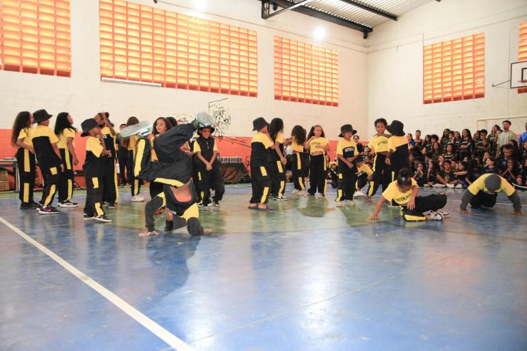 A foto mostra crianças e adolescentes participando de uma apresentação de dança urbana, com movimentos de hip-hop, em uma quadra esportiva coberta. Os participantes vestem uniformes pretos e amarelos, alguns com chapéus, e realizam diferentes movimentos coreográficos, enquanto outros aguardam sua vez. Ao fundo, um grupo de estudantes acompanha a apresentação, sentado e em pé junto à parede.