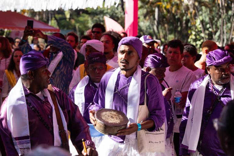 Fotografia em plano médio de um grupo de congadeiros em uma celebração tradicional. No centro, um homem jovem de pele negra, cavanhaque e turbante roxo segura com as duas mãos um pequeno instrumento de percussão metálico (patangome). Ele veste uma camisa roxa adornada com colares de contas e uma faixa branca sobre os ombros. Ao seu redor, outros integrantes do grupo também vestem camisas e turbantes roxos com faixas brancas cruzadas no peito, tocando instrumentos musicais.