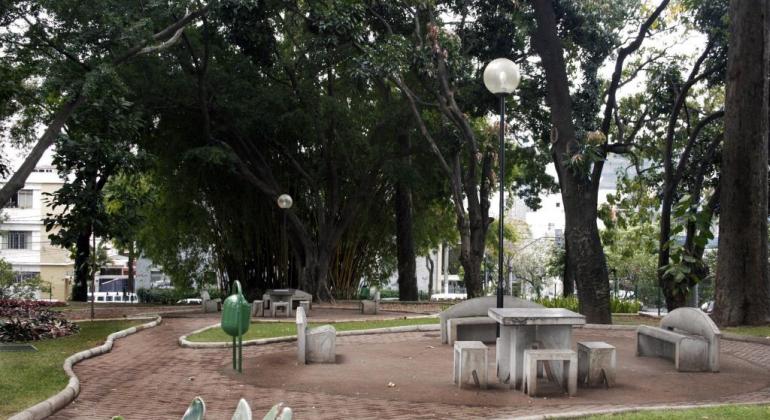Árvores rodeiam calçamento e bancos e mesa de cimento; paisagem do Parque Rosinha Cadar, durante o dia. 