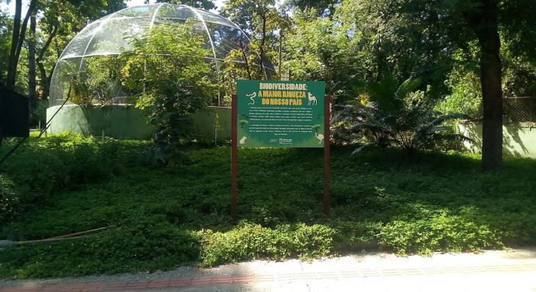Sinalização no zoológico de BH. Árvores e estufa. À frente, placa com os dizeres "Biodiversidade: a maior riqueza do nosso país"