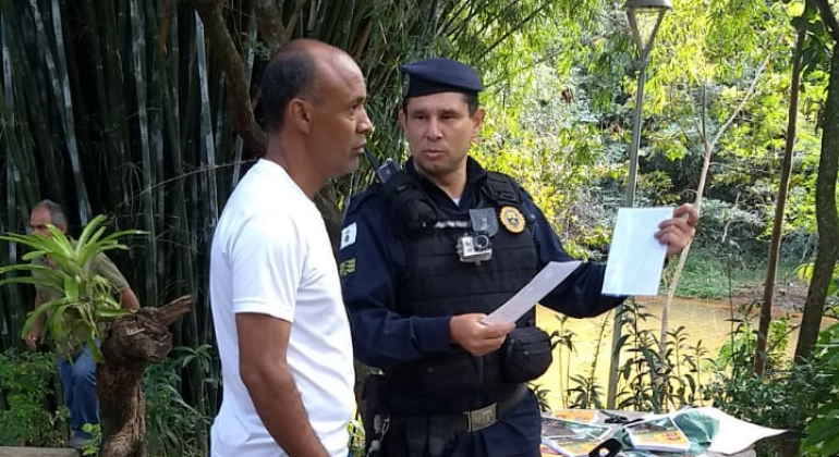 Guarda municipal mostra panfleto a cidadão em parque, durante o dia. 