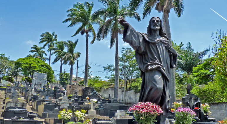 Prefeitura se prepara para receber cerca de 100 mil pessoas nas quatro necrópoles municipais no sábado, dia 2/11. Foto: Divulgação PBH Túmulo com estátua em pedra escura, cercado por outros túmulos e vegetação, durante o dia.