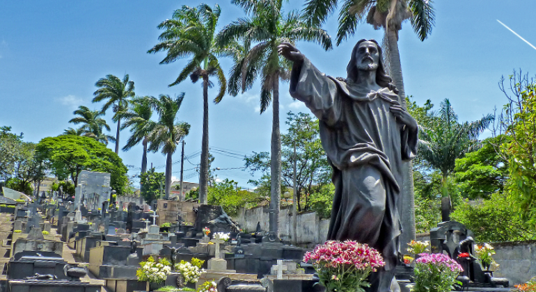 Túmulo com estátua em pedra escura, cercado por outros túmulos e vegetação, durante o dia. 