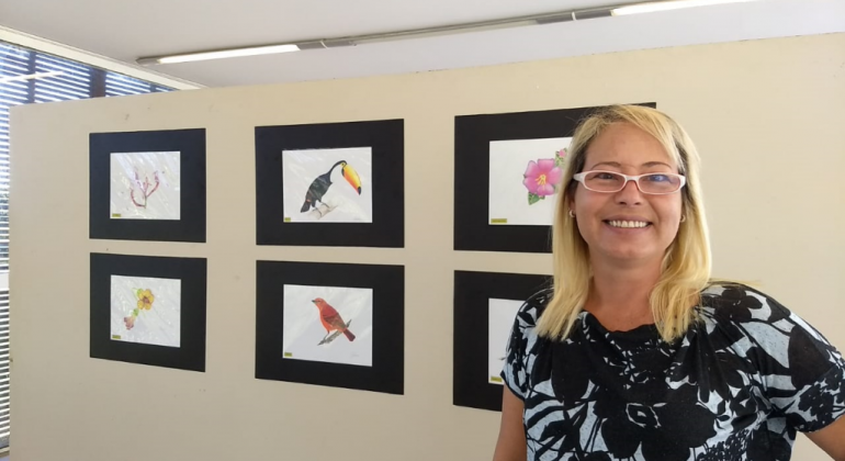 A fauna e flora do cerrado brasileiro são destaques da exposição “Cerrado e Campos Rupestres - Ilustração a lápis de cor”. Foto: Divulgação PBH Mulher sorri, à frente de imagens de pássaros, em exposição.
