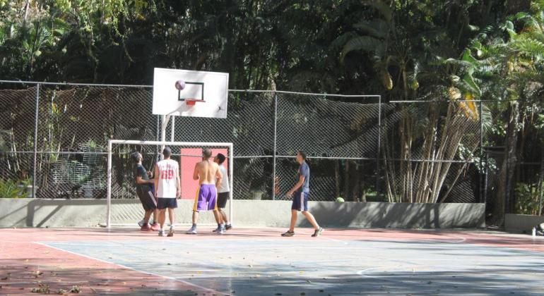 Cinco homens jogam basquete em quadra aberta do Parque Municipal, durante o dia. 