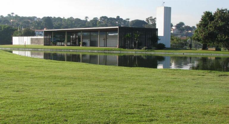 Fachada de construção do Parque Ecológico da Pampulha, com lago e grama à frente, durante o dia. 