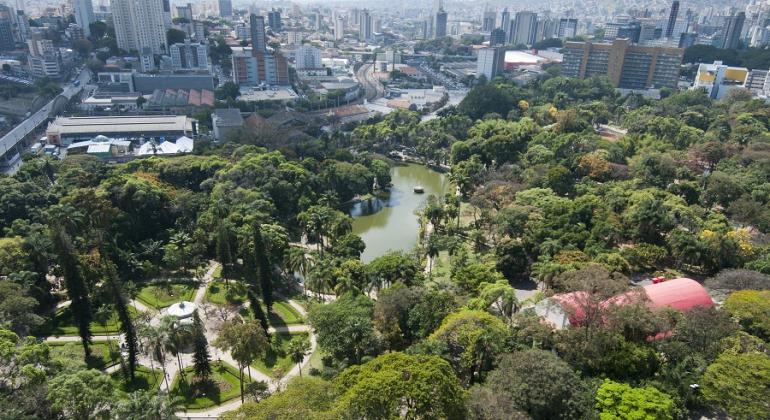 Vista aérea do Parque Municipal, com cidade ao fundo e coreto e lago, durante o dia. 