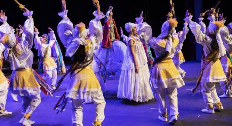 Mulher de vestido e turbante brncos no meio do palco, ao redor dela, homens de roupas brancas e amarelas dançam de maneira coordenada. 