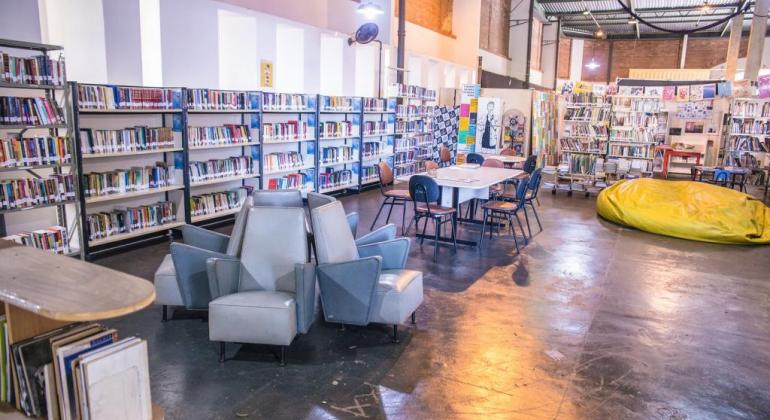 Biblioteca em ambiente amplo, com mais de oito prateleiras, seis sofás, uma mesa com quatro cadeira se um puff