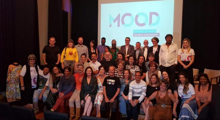 Mais de trinta pessoas, participantes da equipe do MOOd, posam para foto à frente de projeção com a logo do projeto. 