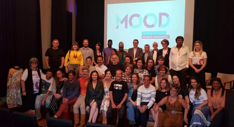 O setor da moda gera atualmente mais de 28 mil empregos diretos e movimenta uma massa salarial de R$ 50 milhões mensais para Belo Horizonte. Foto: Divulgação PBH Mais de trinta pessoas, participantes da equipe do MOOd, posam para foto à frente de projeção com a logo do projeto.