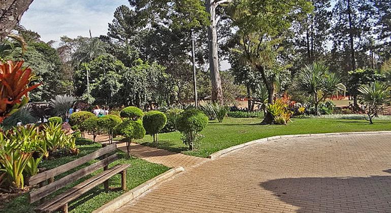 Banco de praça, à esquerda, com vegetação atrás e aos lados; à frente do banco, calçamento; atrás do banco, no alto à esquerda, algumas pessoas passeiam. 