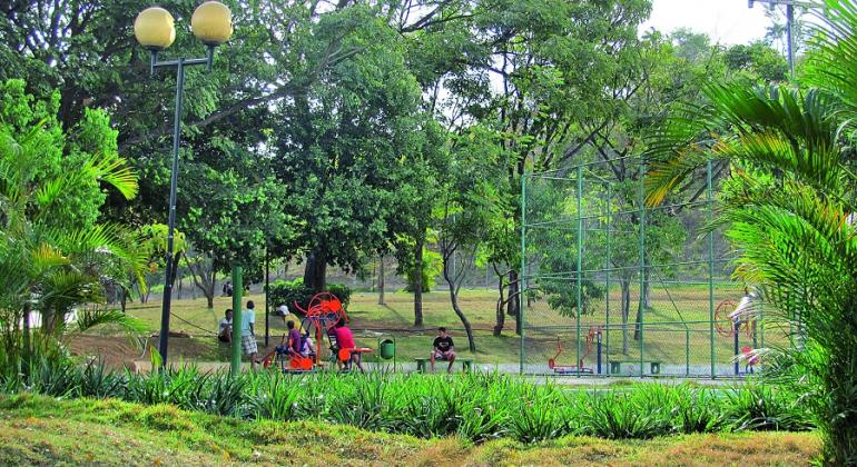 Parque com quadra poliesportiva e aparelhos de ginástica, com pessoas transitando, cercadas de verde, durante o dia. 