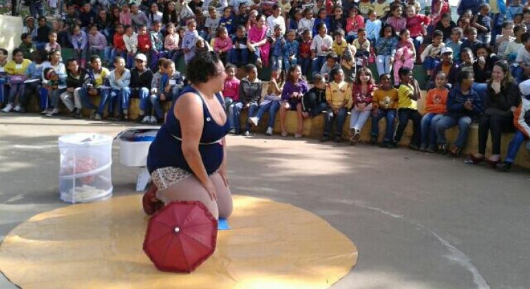 Mulher, vestida de palanha, se apresenta para mais de 50 pessoas sentadas em escadaria circular.