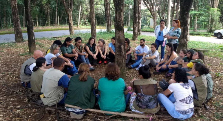 2º Encontro de Monitores Metropolitanos é bimestral e promovido em parceria com o Instituto Estadual de Florestas de Minas Gerais (IEF-MG). Foto: Nadja Simbera Foto: Nadja Simbera Cerca de vinte pessoas, sentadas no chão, em círculo, ao redor de uma árvore, em região arborizada, durante o dia.