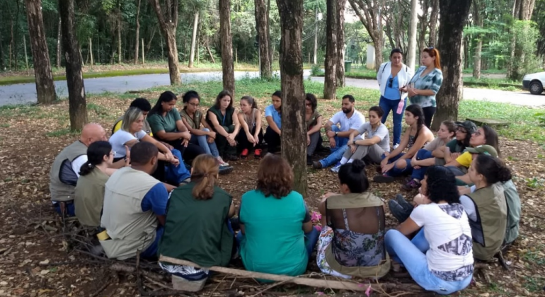 Cerca de vinte pessoas, sentadas no chão, em círculo, ao redor de uma árvore, em região arborizada, durante o dia. 