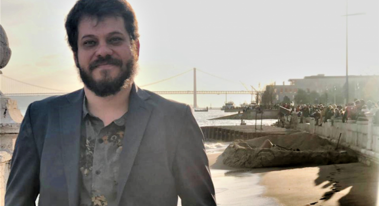 Homem de blazer sorri; no fundo, uma praia e uma ponte, durante o dia. 