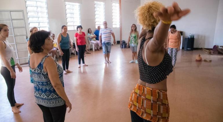 Oito pessoas fazem aula de dança, em uma sala, durante o dia. 