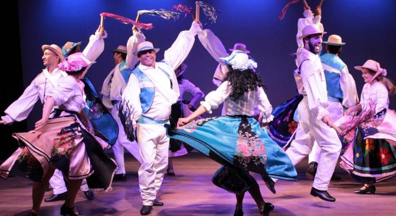 Grupo folclórico Aruanda em apresentação: cerca de cinco casais com roupas folclóricas dançam, em apresentação teatral. 