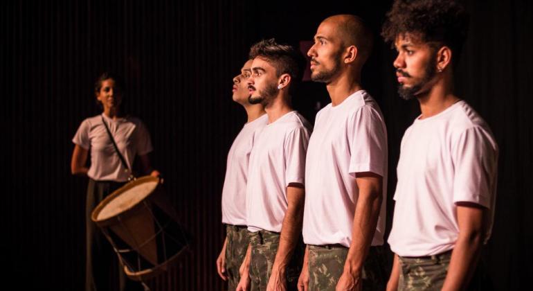 Está em cartaz, nos dias 3 e 4/8, às 20h, espetáculo que investiga situações vividas por gays que passam pelo serviço militar obrigatório. Foto: Gustavo Mazzeti Mulher com tambor ao fundo, à esquerda, e quatro rapazes com camiseta branca e calça militar enfileirados, no centro e à direita.
