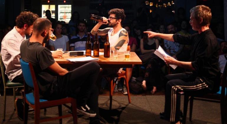 Quatro homens conversam em uma mesa de bar cênica, em um palco