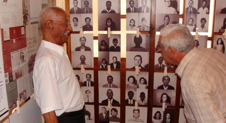 Através do projeto “Cestas da Memória”, idosos voluntários identificam imagens antigas do acervo fotográfico do Arquivo Público. Foto: Acervo PBH Dois homens idosos conferem parede com várias fotos antigas.