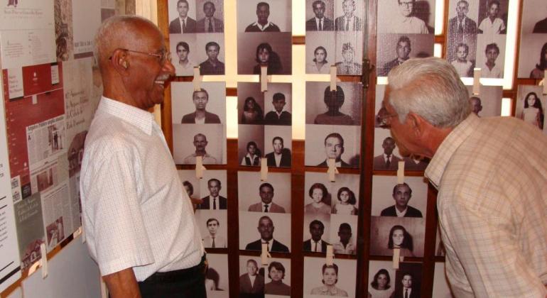 Dois homens idosos conferem parede com várias fotos antigas. 