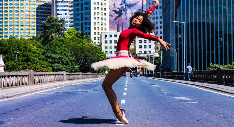 A Fundação Municipal de Cultura traz uma programação sobre o Mês da Consciência Negra em 12 centros culturais da cidade até o dia 24/11. Foto: Márcio Silva Mulher negra com blusa rosa e saia branca de bailarina faz pose de ponta no viaduto Santa Tereza, durante o dia.