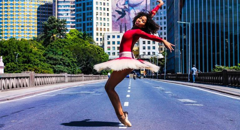 Mulher negra com blusa rosa e saia branca de bailarina faz pose de ponta no viaduto Santa Tereza, durante o dia. 