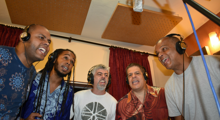 Grupo Agô tem repertório autoral autoral e tem como fonte principal o vasto campo das tradições afro-brasileiras. Foto: Myriam Nacif Cinco homens, em estúdio, cantando.