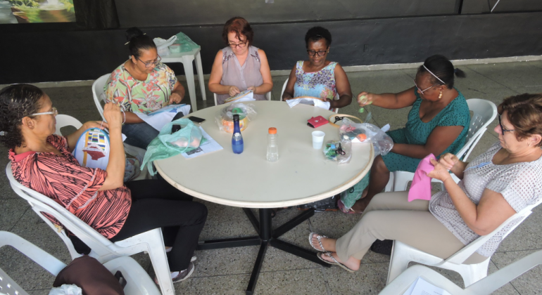 Cinco mulheres bordam, cada uma sua peça, todas sentadas em torno de uma mesa. 