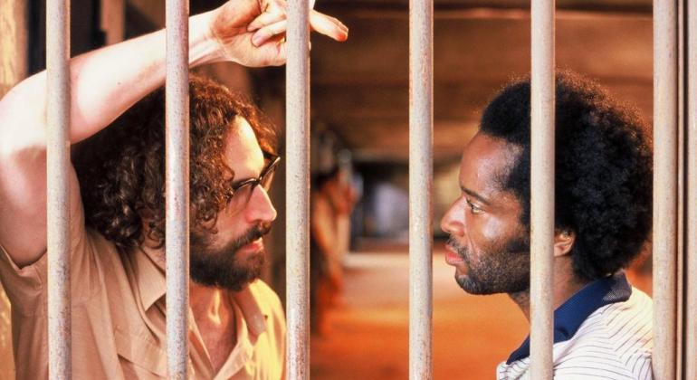 Fundação Municipal de Cultura apresenta na terça, dia 18/12, às 19h30, no MIS Cine Santa Tereza, o projeto “Diálogos MIS”, com Paulo Lins. Foto: Divulgação Homem branco, barbudo e com cigarro na mão olha para homem negro; ambos estão atrás das grades. Cena do filme "Quase dois irmãos".