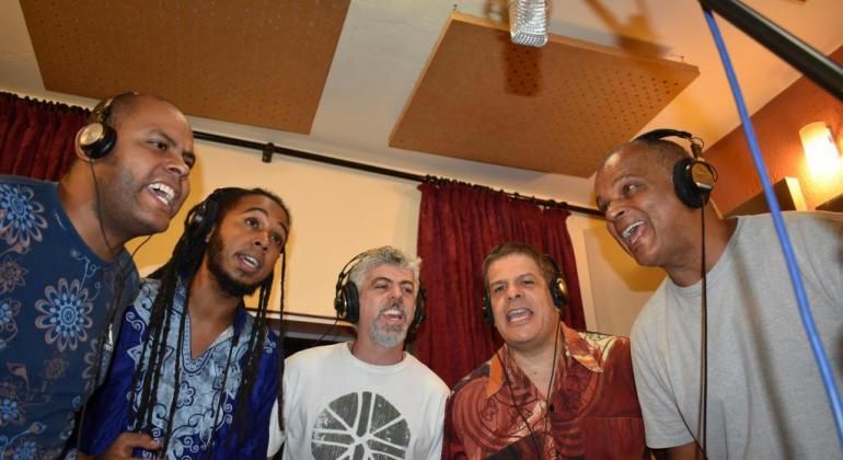 Cinco homens, em estúdio, cantam. 