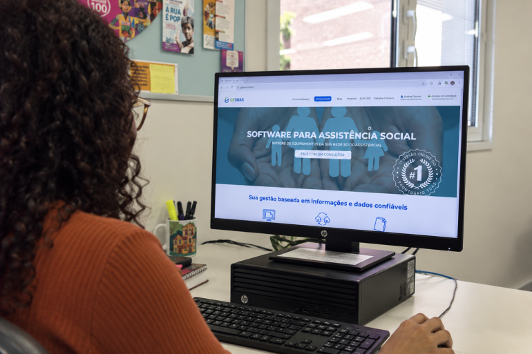 Prefeitura implementa nova ferramenta para integrar dados e atendimentos da Assistência Social Uma mulher de cabelos longos e cacheados, vista de costas, está sentada diante de um computador. Ela usa óculos, blusa marrom e mantém uma das mãos sobre o mouse. Na tela do computador está aberta a página do software Gesuas. Sobre a mesa branca há teclado, mouse, computador e um recipiente com canetas. Ao fundo, há uma janela que ilumina o espaço com luz natural e um mural com informativos. O ambiente tem aparência de eescritório, com organização simples e cotidiana.