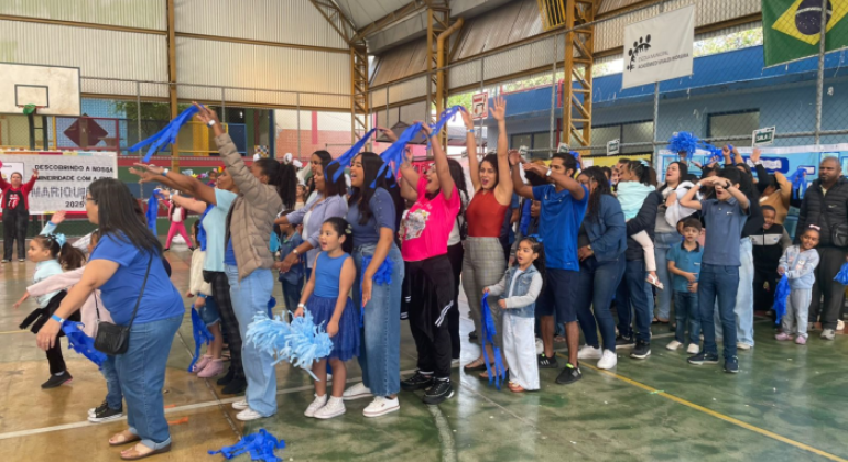 #paratodosverem: a imagem mostra um grupo animado de crianças e adultos participando de uma gincana em uma quadra esportiva. Alguns(mas) levantam os braços e seguram pompons e fitas azuis. O ambiente está decorado com faixas e a bandeira do Brasil. Ao fundo, uma faixa diz "Descobrindo a nossa mineiridade com a Emei Mariquinhas – 2025".