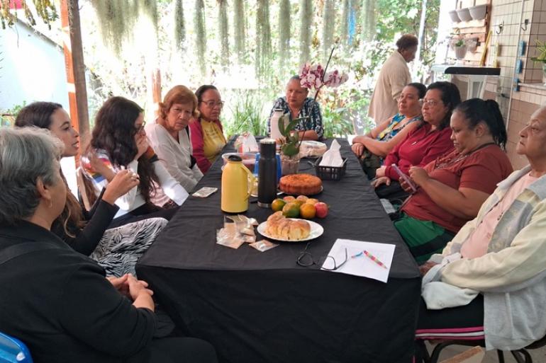 Grupo de alimentação saudável transforma orientações em cuidado coletivo no Centro de Saúde Alcides Lins A imagem, em ambiente externo, mostra um grupo de cerca de dez mulheres reunidas ao redor de uma mesa, participando de reunião acompanhada de um lanche com bolo, pães, frutas e garrafas térmicas.