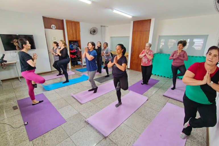 Prática de yoga favorece a mobilidade e promove o bem-estar aos atendidos no Creab Centro-Sul Onze mulheres em uma sala com tapetes coloridos em pé fazendo aula de yoga.