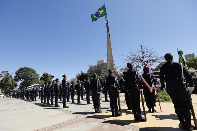 Guardas municipais de Belo Horizonte vestem uniforme azul marinho e estão alinhados em formação durante uma cerimônia ao ar livre na praça da Bandeira. No centro, uma bandeira do Brasil tremula no alto de uma haste sob o céu limpo. 