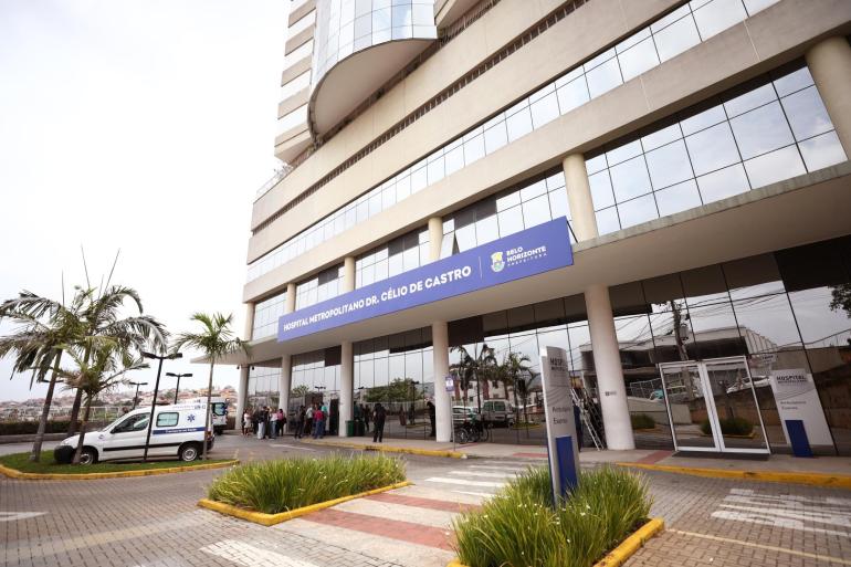 A imagem mostra a fachada do Hospital Metropolitano Dr. Célio de Castro. A foto contém também imagens de palmeiras e uma ambulância estacionada