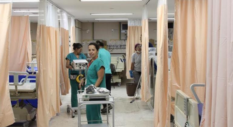 TRês profissionais de saúde, com carrinhos e material hospitalar, carregam equipamentos em meio a macas de hospital, em sala do Hospital Metropolitano Odilon Behrens. 