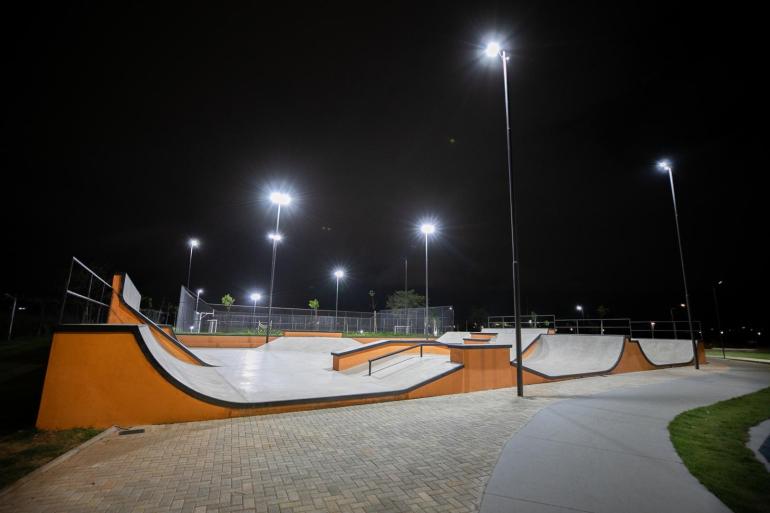 Visão noturna de uma pista de skate ao ar livre, vazia, com rampas de concreto cinza e detalhes laranjas. Vários postes altos iluminam fortemente o local