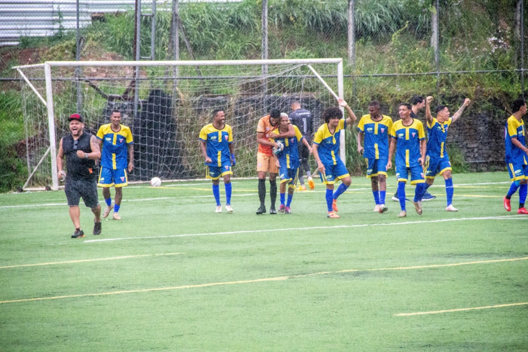 Jogadores da Copa Centenário, usando uniformes azul e amarelo, comemoram uma jogada em campo, enquanto companheiros e integrantes da equipe celebram ao fundo.