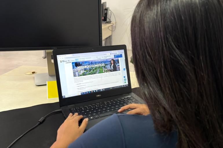 PBH oferece curso gratuito para ampliar conscientização sobre mudanças climáticas Mulher de cabelos escuros, vista de costas, utiliza um notebook em um ambiente de trabalho. Na tela, está aberta uma página do site da Prefeitura de Belo Horizonte com informações sobre mudanças climáticas.