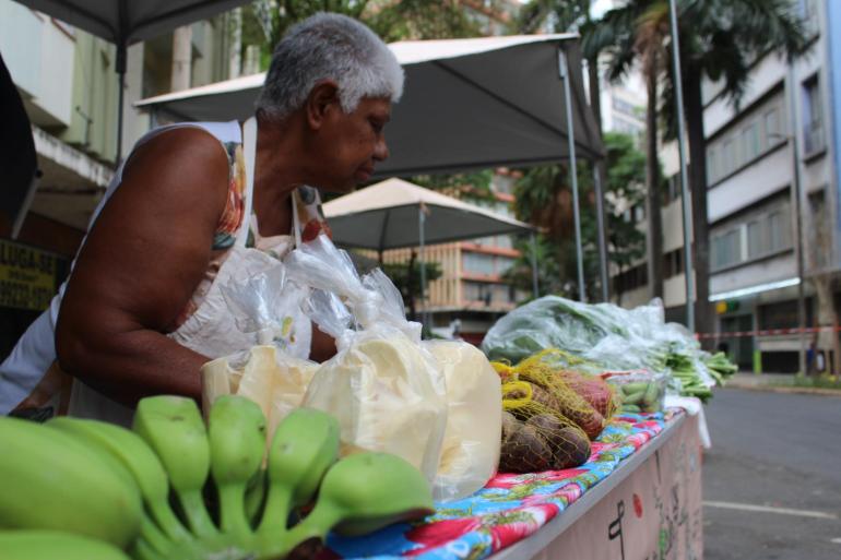 Mulher negra expõe bananas verdes e maduras em uma tenda de frutas, organizadas sobre a banca para comercialização